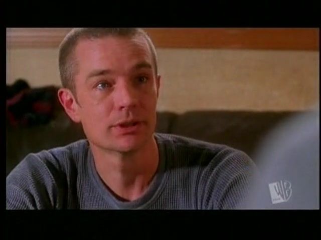 The image shows a man seated indoors, looking slightly to his side. He has short, light-colored hair and is wearing a textured, long-sleeve gray shirt. In the background, there is a blurred brown couch and a neutral wall. The lighting is soft, creating a warm atmosphere. The bottom right corner features a small logo with the letters "WB."