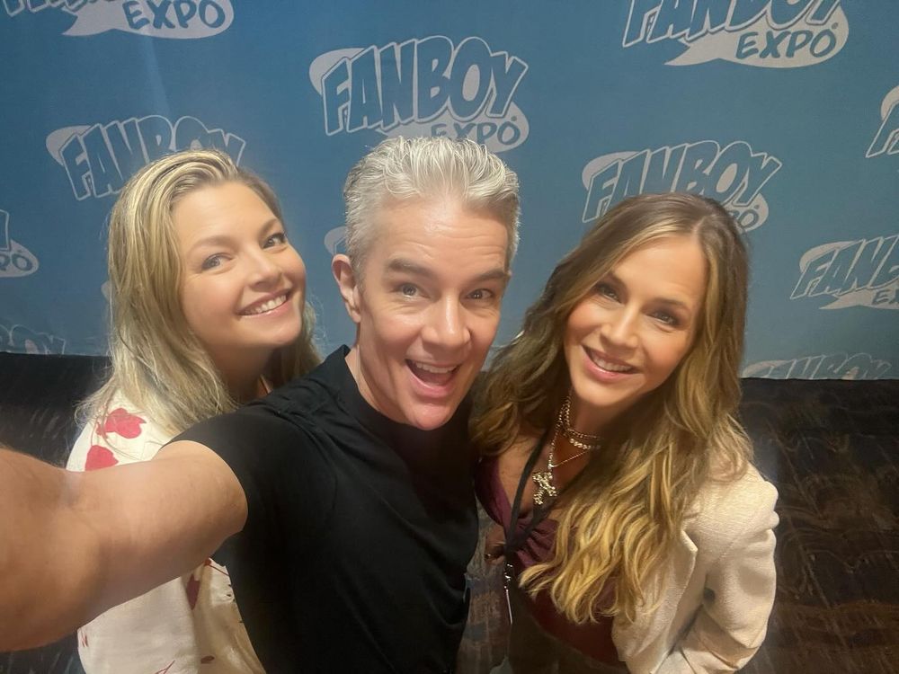 The image shows three individuals posing together for a selfie at what appears to be a convention or expo. The person in the center is taking the selfie with an outstretched arm. They have short, light hair and are wearing a black T-shirt. On the left is a person with long blonde hair wearing a white shirt with red patterns. On the right is another person with long, wavy hair wearing a beige jacket over a maroon top, accessorized with a necklace. The background consists of a blue wall with repeated white text logos that read "FANBOY EXPO."