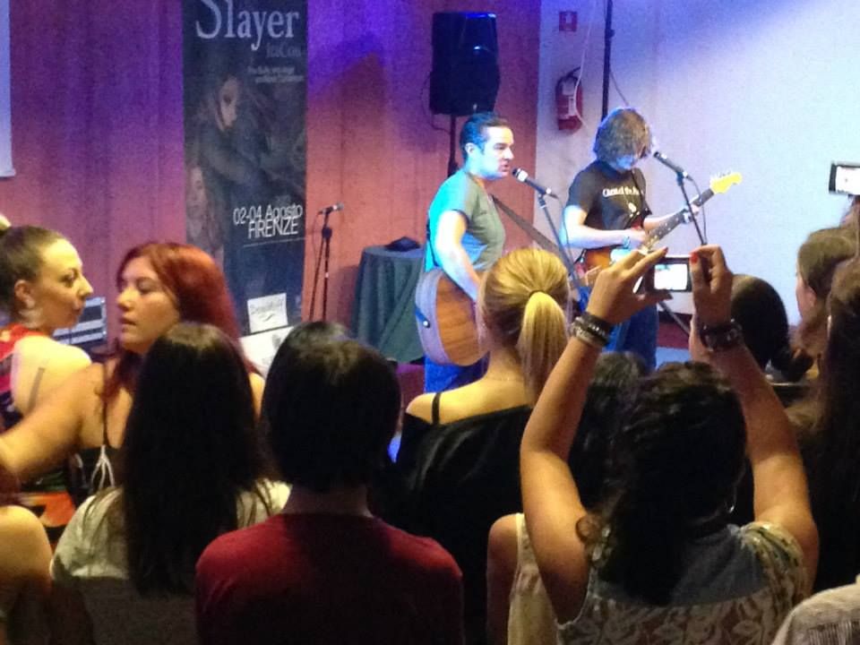 The image shows a small live music performance in what appears to be an intimate indoor venue. Two musicians are on stage, both playing guitars and positioned in front of microphones. The musician on the left (James) is playing an acoustic guitar, while the one on the right (Sullivan) has an electric guitar. A group of people stands in front of the stage, closely packed together, watching the performance. The audience's attention is focused on the musicians, with one person holding a smartphone up to take photos or record. The background features a wooden wall with a poster visible on the left side. The lighting creates a warm and casual atmosphere, with soft, diffused light illuminating the scene.