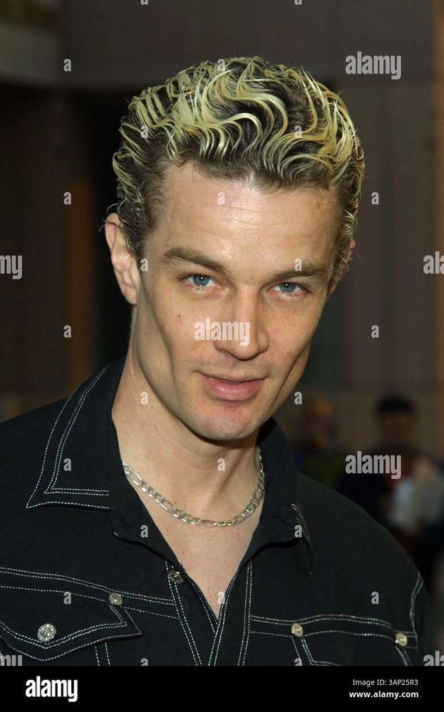 The image depicts a person with short, curly hair styled upward, featuring bleached blonde highlights. The individual is wearing a dark shirt with detailed white stitching and a chain necklace. The background is out of focus, suggesting an indoor setting with low lighting.