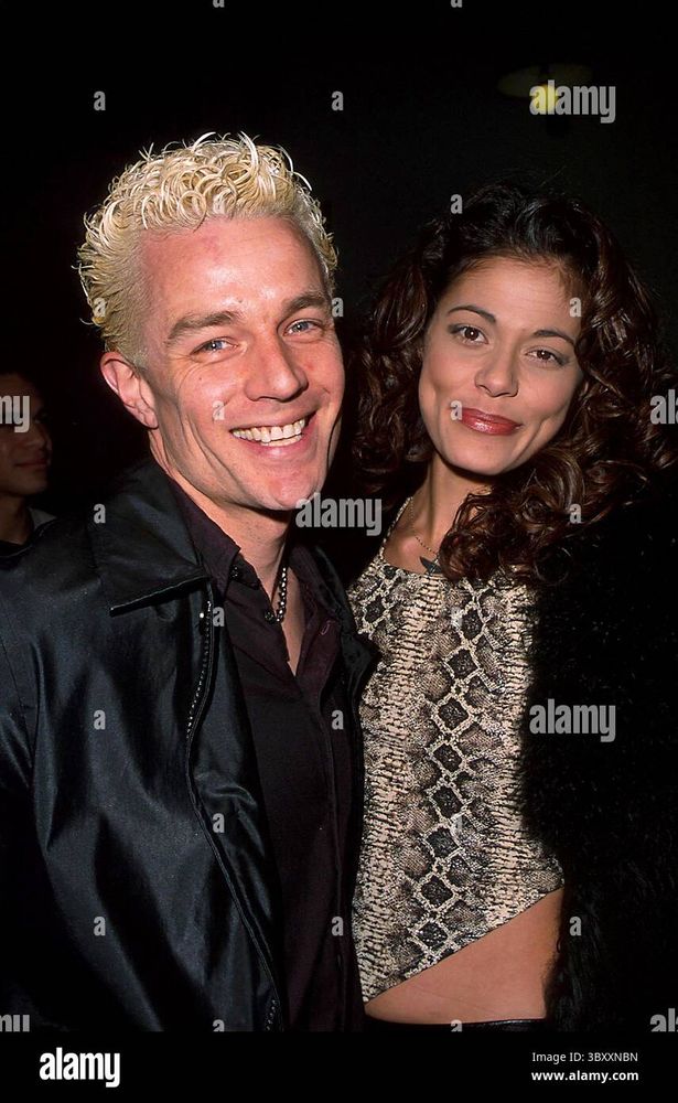 The image shows two individuals posing closely together. On the left, a person with short, curly blonde hair is wearing a black leather jacket over a dark shirt. The individual has a broad smile and is slightly leaning towards the person next to them. On the right, another person with long, dark, curly hair is wearing a textured, patterned top resembling snakeskin under a black, fuzzy jacket. Both appear to be in a dimly lit environment, possibly a social or formal event.