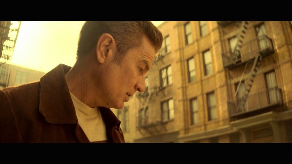 The image portrays a side profile of a person with short hair outdoors, looking downward. The background consists of a brick building with several windows and external fire escapes, typical of urban architecture. The lighting has a yellowish tint, suggesting a sunset or sunrise, which casts a warm hue over the scene. The person wears a brown jacket over a light-colored shirt. The composition focuses on the person in the foreground, with the building softly blurred out in the background.