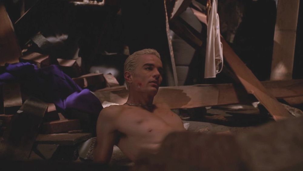 The image shows a shirtless man with short, bleached hair leaning against debris in a dimly lit setting. He appears to be in a disarrayed environment with scattered wooden planks and draped fabric. The lighting casts shadows, creating a moody atmosphere. Some structural elements like beams and fabric can be seen in the background.