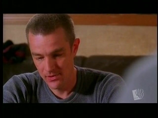 The image shows a person with short hair and a focused expression seated indoors. They are wearing a textured, gray sweater and seem to be engaged in a conversation or deep thought. The background is softly blurred, indicating a cozy interior setting. In the bottom right corner, a small logo is visible.