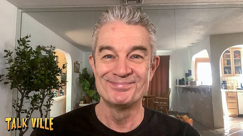The image shows a person smiling at the camera, set against a domestic background. The individual has short gray hair and is wearing a black shirt. Behind the person is a room with bright lighting, featuring furnishings such as a brown curtain, a potted plant, and a dining area with chairs. The background includes a kitchen area with open shelves and various kitchen items. The walls are light in color, accented by an archway leading to another part of the house. On the bottom left of the image, the text "TALK VILLE" is visible in yellow uppercase letters.