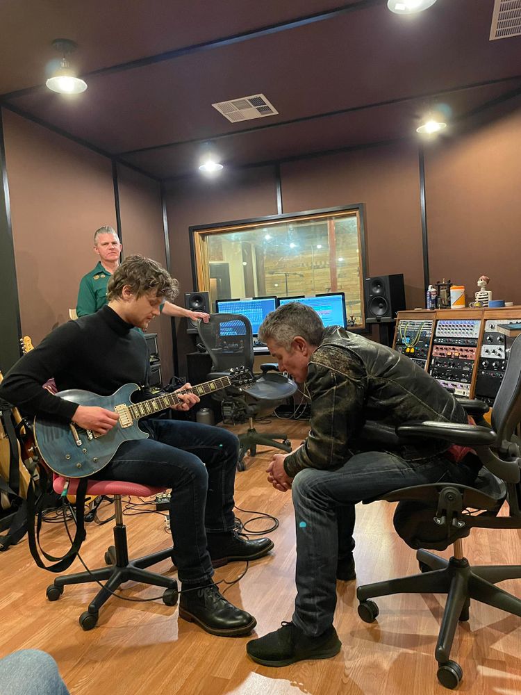 The image shows a recording studio with three people present. In the foreground, a person is playing a blue electric guitar while seated on a swivel chair. They wear a black turtleneck and dark pants. Next to them, another person, wearing a leather jacket and jeans, leans forward, listening attentively. In the background, a third person stands near a mixing console, observing. The studio has wooden floors, and sound panels line the walls. Equipment, including speakers, monitors, and a mixing board with colorful knobs, is visible. There is a window into another room with a view of the studio's interior.