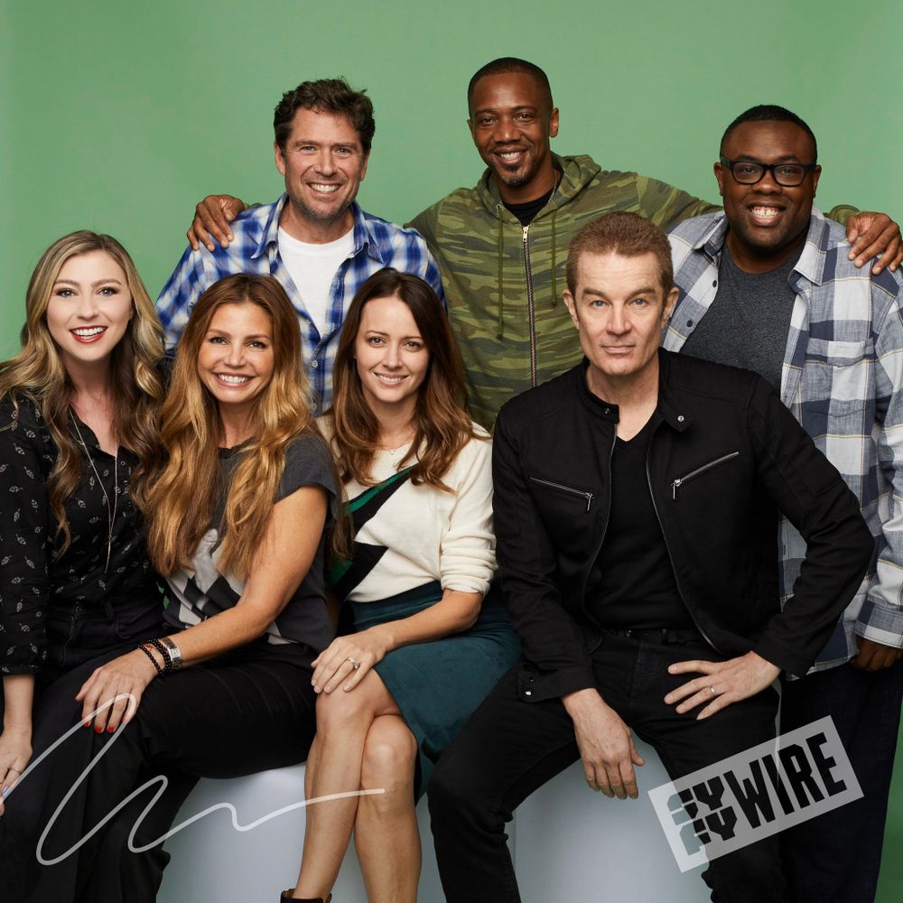 The image features a group of seven people sitting and standing together against a solid green background. Four people are seated in the front, while three are standing behind them. The individuals display friendly and approachable expressions. The person on the left wears a black patterned blouse and has long, light-colored hair. Next to her is a woman with long, wavy, reddish-brown hair, wearing a dark casual top. The third seated person has medium-length brown hair and wears a white sweater with a dark green pattern. The last seated person on the right has short, light hair and wears a black jacket with zippered pockets. Behind them, the person on the far left stands in a blue plaid shirt with a hand on the shoulder of the seated women. In the middle stands a person in a green camouflage jacket, and the person on the far right wears a plaid shirt with glasses. The image features two logo watermarks in the bottom corners.