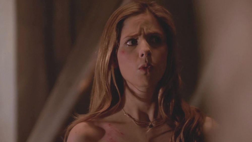 The image shows a person with long, light brown hair looking slightly to the side with a concerned expression. The lighting is dim, casting shadows on their face. They have some scratches visible on their shoulder. The background is out of focus, creating a sense of depth with blurred shapes in neutral tones.