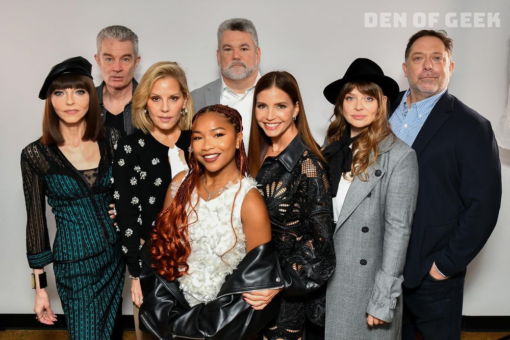 The image features a group of eight people posing together in front of a plain white background. The individuals are arranged in two rows. In the back row, three men and two women are standing. They are dressed in a mix of semi-formal and stylish outfits, including a gray blazer, black and white clothing, and a hat. In the front row, three women are positioned slightly forward. They wear a variety of textured and patterned garments including a black leather jacket, a lace dress, and a textured white top. The group appears cheerful, looking at the camera with smiles and relaxed expressions.