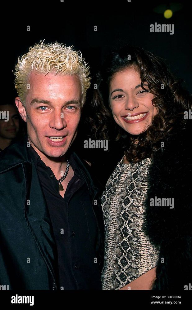 The image features a man and a woman posing closely together at an indoor event. The man on the left has short, curly, bleached blonde hair and is wearing a black shirt with a dark jacket. He has a silver necklace with a pendant. The woman on the right has long, wavy, dark brown hair and is smiling brightly. She is wearing a patterned top with a black coat. The background is dark, suggesting a nighttime setting, with other people and dim lights faintly visible. The watermark "alamy" is repeated across the image.