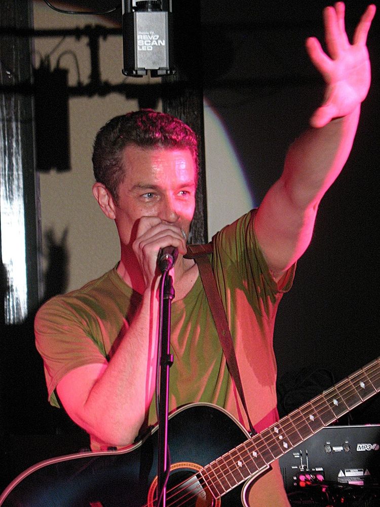 The image shows a person performing on stage, holding a microphone close to their mouth using their left hand while their right arm is raised. They're wearing a casual olive-green t-shirt and stand behind a black acoustic guitar. The lighting casts a pink hue over the scene, creating a vibrant atmosphere. Above, a light fixture labeled "Revo Scan LED" is visible. The background is dark with soft shadows.