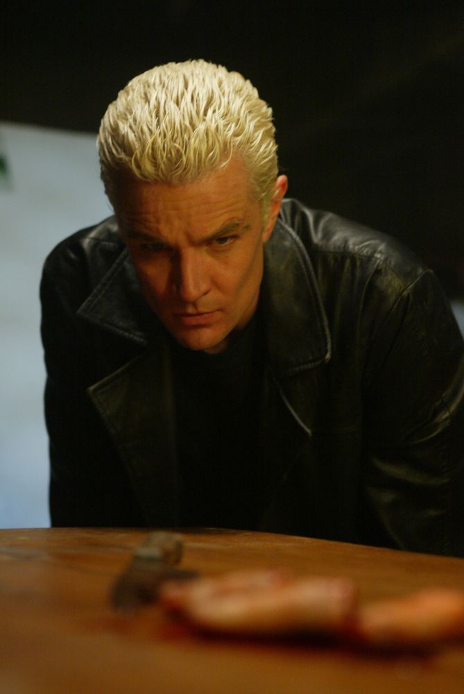 The image depicts a person with short, bleached blond hair leaning forward slightly, with an intense expression. The individual is wearing a black leather jacket over a dark shirt. The background is dimly lit, creating a moody atmosphere. In the foreground, there's a blurred object, possibly on a table, making it unclear what it is.