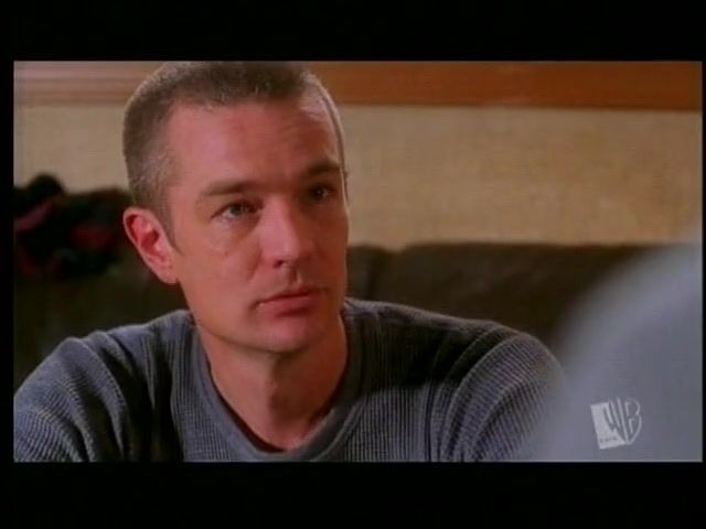 The image shows a close-up of a person with short hair, looking intently towards the left side of the frame. They are wearing a gray ribbed sweater. The background is out of focus, showing neutral, muted tones with what appears to be a dark sofa or cushion and a wooden or beige wall.