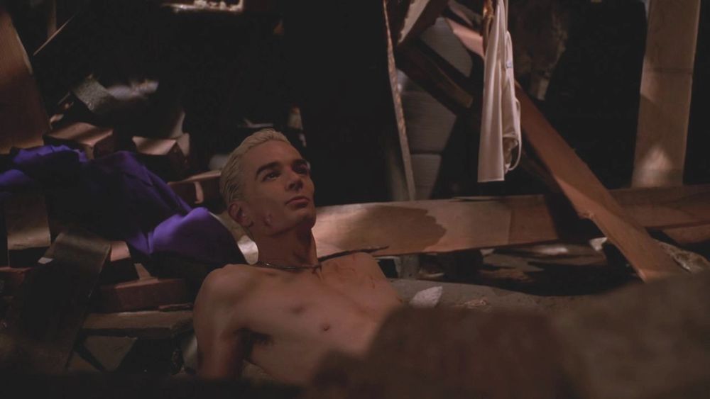 The image shows a shirtless man with short, light-colored hair reclining in a dimly lit, cluttered space. He appears relaxed, leaning back slightly with his head resting against some wooden beams. The background is filled with various debris, including wooden planks and possibly fabric, suggesting an attic or another storage area. A piece of light-colored cloth hangs in the upper right corner.