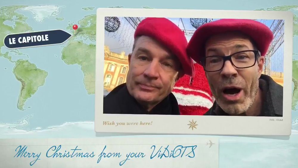 The image is a holiday-themed postcard featuring two people wearing red berets. They are positioned in the foreground, with one person on the left and the other on the right. Both appear to be looking directly at the camera. The person on the right is wearing glasses. They stand in front of a festive background with hanging decorative lights and an ornate building in the distance. A cream-colored border surrounds the photo, emulating a classic postcard style. In the background, a world map is visible with a pin pointing at a location labeled "Le Capitole." Below the image, in a script font, is a holiday greeting.