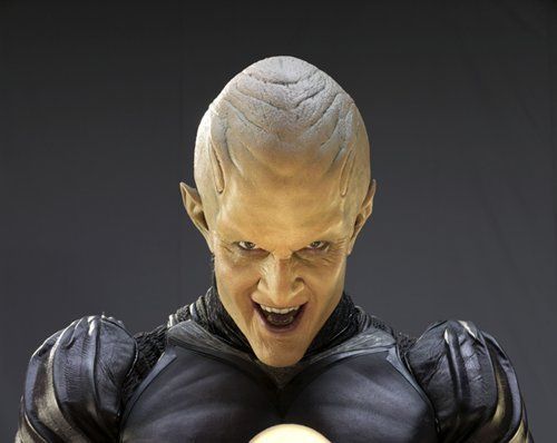 The image shows a humanoid character with an exaggeratedly smooth and elongated bald head, featuring pronounced ridges on the forehead. The skin appears textured and is a pale, muted color. The character has pointed ears that add to its unique appearance. Their expression is intense and features slightly parted lips showcasing a menacing look. The character is dressed in a form-fitting, dark-colored suit with padded shoulders and detailed fabric texture.