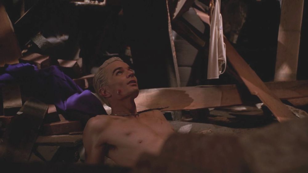 The image shows a shirtless man with blonde hair lying on a wooden floor amidst debris and broken wooden beams. The lighting is dim, casting shadows across the scene, with hints of a purple cloth nearby. The man is looking upwards, appearing to be in a contemplative or pained state. In the background, an article of light-colored clothing hangs from a beam. The setting suggests a collapsed or damaged indoor environment with scattered objects and a dark, cluttered backdrop.