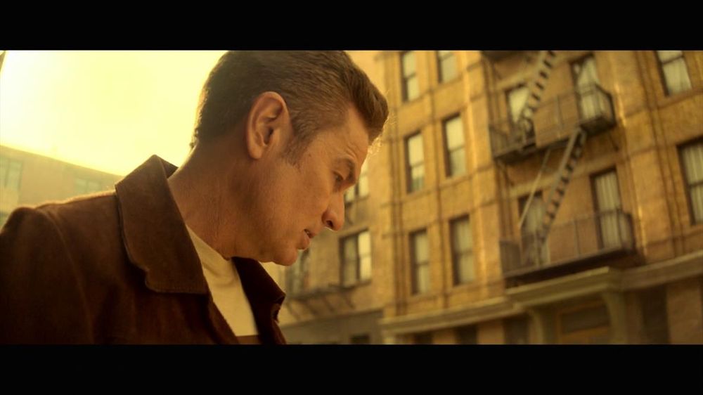 The image shows a man in profile, looking downward, positioned on the left side of the frame. He is wearing a brown jacket over a light-colored shirt. His hair is short and neatly combed. The background features a yellowish tint, indicating a warm or sunset atmosphere. A multi-story brick building with multiple windows and an external fire escape ladder occupies the right side of the frame. The building's facade suggests an urban setting.