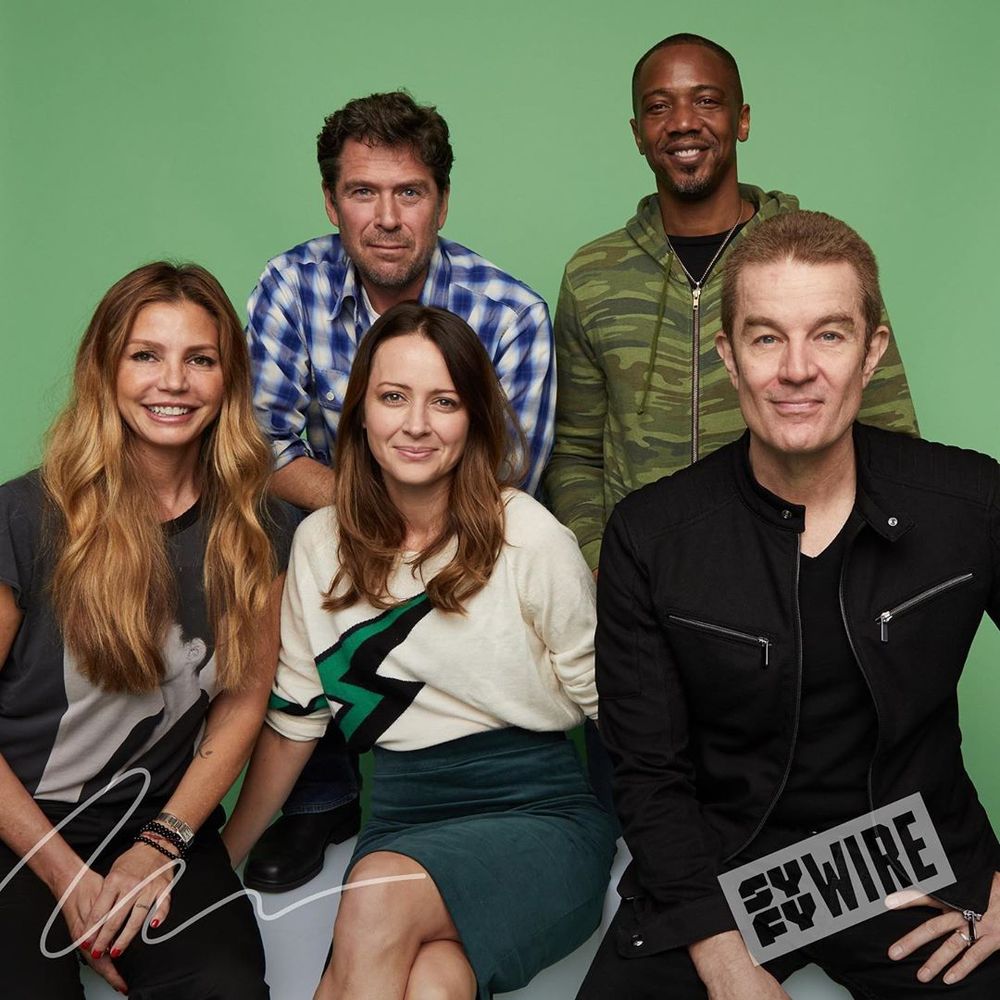 The image shows a group of five adults posing against a solid light green background. They are arranged in two rows. In the front row, three people are seated. On the left, a woman with long, wavy hair wears a black T-shirt with a graphic design. In the middle, a woman with shoulder-length hair wears a cream sweater with a black and green design. On the right, a man with short hair wears a black jacket with zipper details. Behind them, two men stand. The man on the left behind wears a blue and white checkered shirt, and the man on the right wears a camouflage hoodie. A small "SYFY WIRE" logo is visible at the bottom right corner of the image.