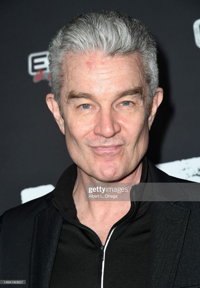 The image shows a close-up of a person with short, gray hair and a slight smile. They are wearing a black jacket with a zipper visible at the collar, set against a dark background. The expression is calm and the lighting highlights the features clearly.