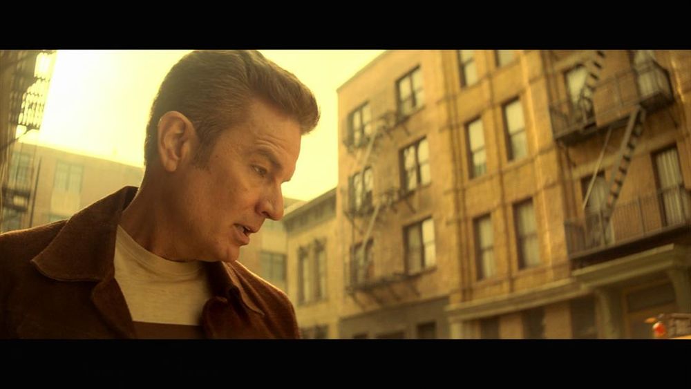 The image portrays a side profile of a person in an urban setting, bathed in warm, golden sunlight. The individual is wearing a brown jacket over a light-colored shirt and is gazing intently ahead. In the background, there are brick buildings with multiple windows, typical of a cityscape. The buildings have external fire escape ladders. The overall tone is warm, with a noticeable yellow tint that suggests either a sunset or sunrise.