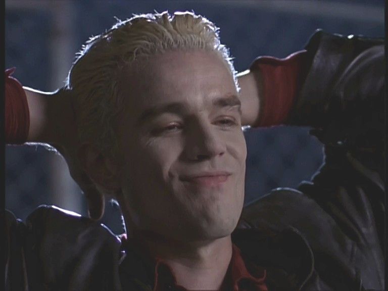 The image depicts a person with bleached blonde hair, styled back smoothly, against a softly focused background. The individual is wearing a dark leather jacket over a bright red shirt. They have their arms raised behind their head in a relaxed posture and are giving a slight, confident smile. The lighting emphasizes the contours of their face and the details in the clothing, creating a juxtaposition of shadows on the skin and jacket.