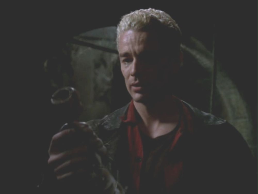 The image shows a person with short, bleached blonde hair standing in a dimly lit environment, possibly a cave or underground setting. The person is wearing a dark leather jacket over a dark shirt with a red collar. They are holding a cylindrical object with both hands, inspecting it closely. The background is blurred and shadowy, with only faint outlines of the surroundings visible, enhancing the mysterious atmosphere.