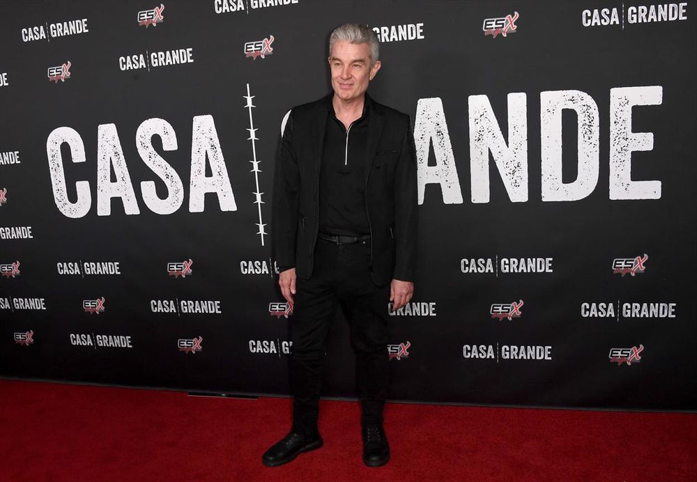 A person stands on a red carpet in front of a black backdrop. The backdrop features the words "CASA GRANDE" repeatedly in large, white, distressed lettering. The words are spaced equally on either side of a white vertical line. Smaller logos reading "ESX" in red and white are also visible across the backdrop. The individual is wearing an all-black outfit, including a jacket, pants, and shoes, and stands with a relaxed posture.