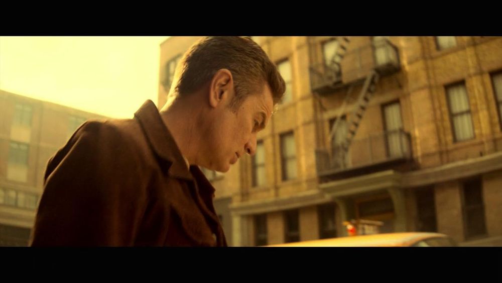 The image portrays a man in profile view on a city street, bathed in warm, golden sunlight. He is wearing a brown jacket and has short, brown hair. In the background, a multi-story brick building with multiple windows and a fire escape ladder is visible. The building has a weathered appearance, consistent with older urban architecture. The image has a soft, vintage filter with strong backlighting from the sun, creating a nostalgic atmosphere.
