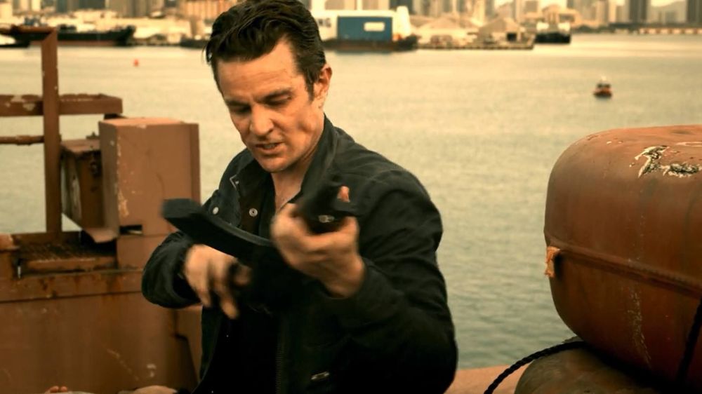 The image depicts a person standing on a rusty metal structure by the water, holding a black object resembling a weapon. The individual is wearing a dark jacket and has short, dark hair. In the background, an urban skyline with various buildings is visible across the water. The surface of the water appears calm, with a few distant boats. The lighting suggests late afternoon or early evening, with a warm hue cast over the scene.