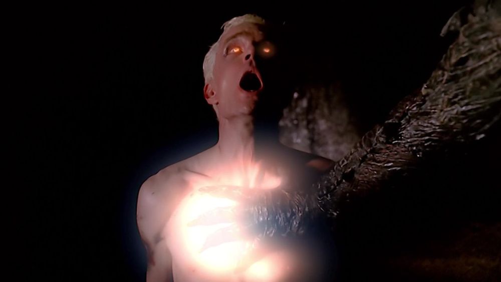 The image depicts a dramatic scene featuring a person with an open mouth, looking upwards. The individual appears to be in distress or awe. A bright, glowing light emanates from their chest area, illuminating their upper body. A dark, textured arm or appendage reaches towards the glowing area, its surface rugged and possibly scaly. The background is dark, highlighting the contrast between the bright light and the surrounding darkness.