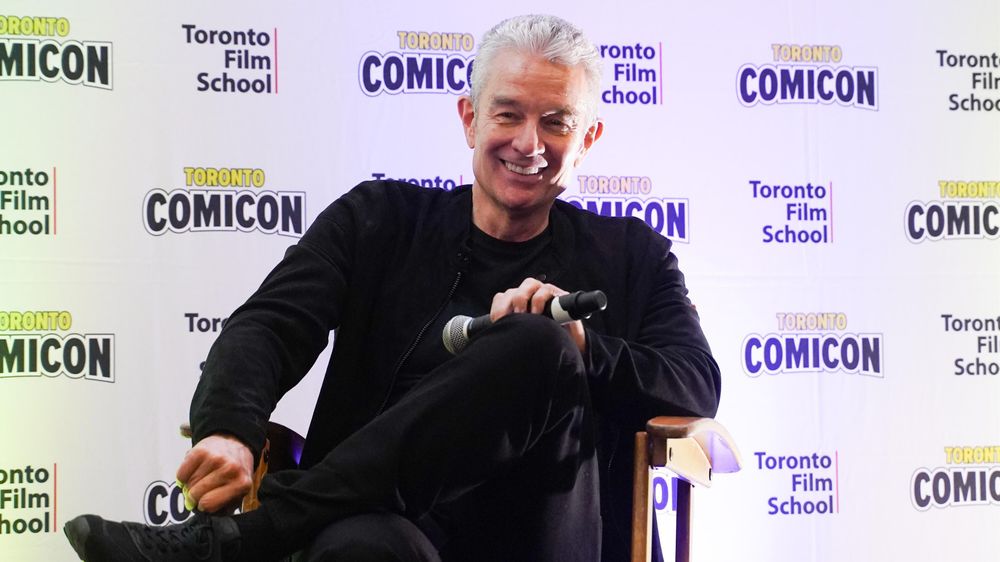 The image shows a man sitting on a chair holding a microphone. He is wearing a black jacket and crossing his legs. The backdrop features repeated logos for "Toronto Film School" and "Toronto ComicCon." The man is smiling and appears relaxed, suggesting he might be participating in a panel or discussion at the event.