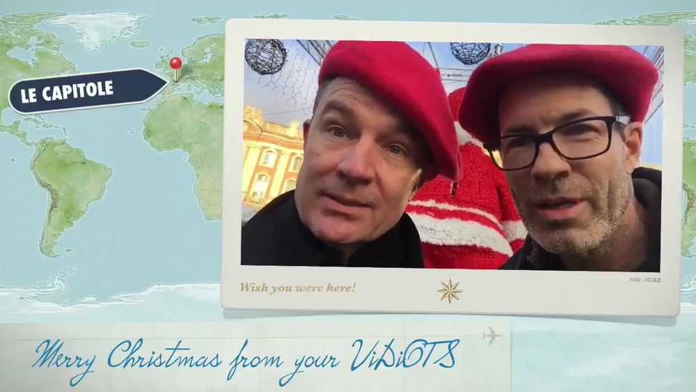The image is a postcard-style composition featuring a map in the background and a photograph in the foreground. The map shows a portion of the world, with a red marker pointing to an area labeled "LE CAPITOLE" in a black label, suggesting a travel or location theme. The photograph shows two men wearing red berets. The man on the left is looking directly into the camera, while the man on the right, wearing glasses, is slightly leaning in. Behind them is a decorative background including wireframe spheres and buildings. Above the photo is a stylized message in a script font that reads "Wish you were here!" followed by the number "NO. 1032." At the bottom of the image, written in blue cursive is "Merry Christmas from your ViDiOTS."