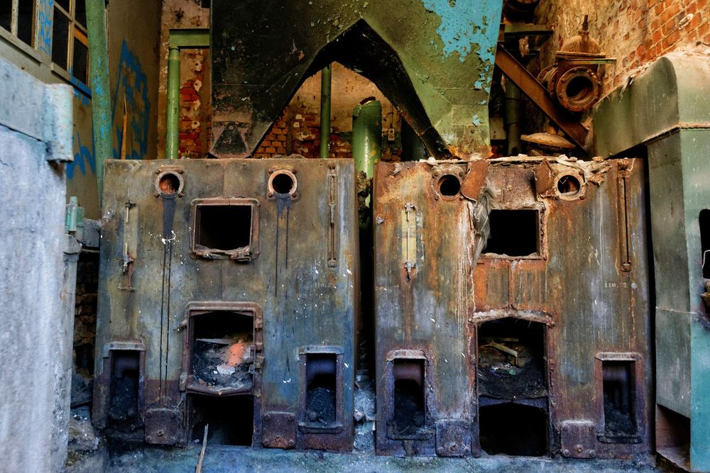 Das Bild zeigt zwei große, verrostete Industrieöfen oder Heizkessel in einem verlassenen Gebäude. Die Metallfronten sind stark gealtert, mit Rost, Abplatzungen und dunklen Verfärbungen. Jede Einheit hat mehrere Öffnungen: runde Luken oben, rechteckige Türen in der Mitte und größere Öffnungen unten, teils mit Ruß und Asche. Die Umgebung wirkt heruntergekommen, mit bröckelndem Putz, freiliegenden Ziegelwänden und grünen Rohren im Hintergrund. Oberhalb der Kessel ist eine große, grün lackierte Metallkonstruktion zu sehen, deren Farbe ebenfalls abblättert. Rechts und links stehen weitere alte Maschinen- oder Rohrbauteile.
