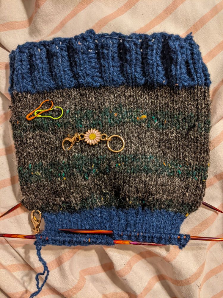 Photograph of a partially finished knitted sock, cast onto double-pointed needles. The cuff and leg have been finished, and the heel flap has been partially completed. There are a few stitch markers connected to the leg of the sock. End image ID.