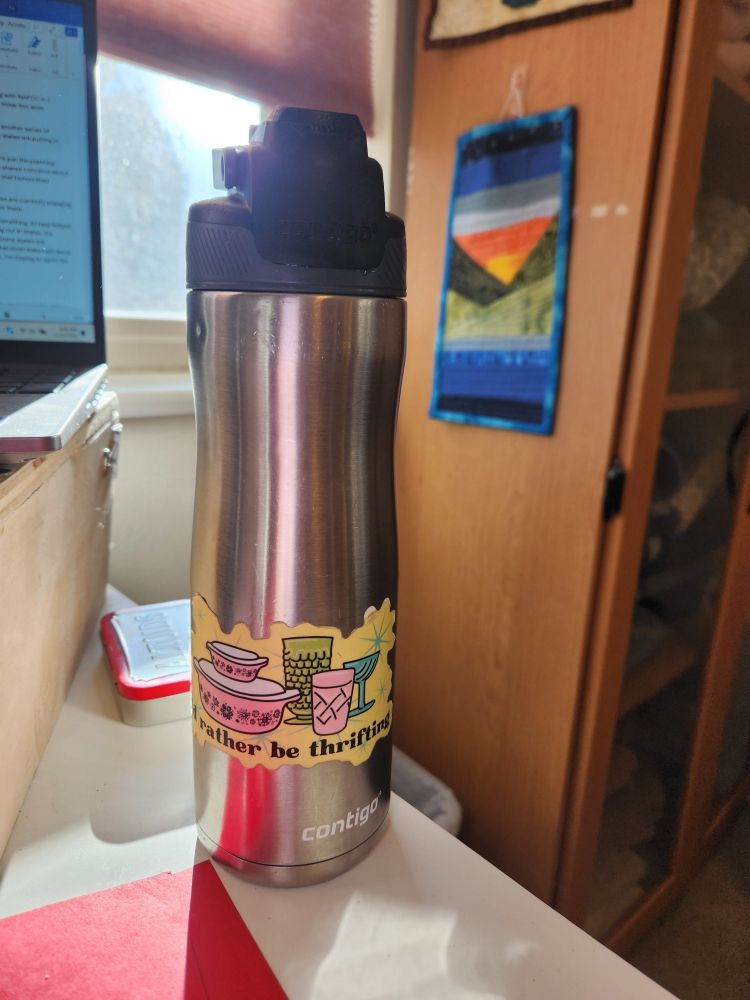 A silver water bottle with a sticker that says, "I'd rather be thrifting." 
Background is a part of a desk, computer, window, and quilted wall hanging.