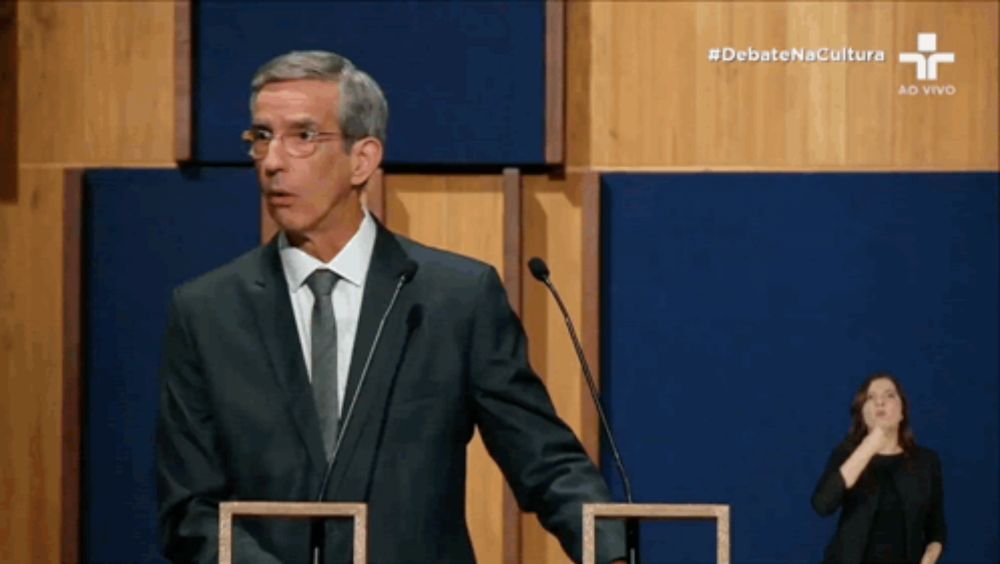 a man in a suit and tie stands at a podium in front of two microphones with debate na cultura written in the corner