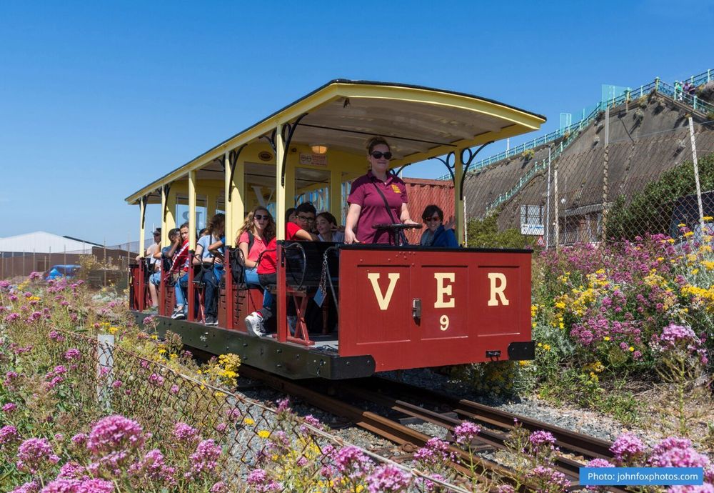 Taken from behind a low mesh fence surrounded by wildflowers, a red & yellow VER car travels towards camera with the driver and passengers looking to camera on a bright sunny day.