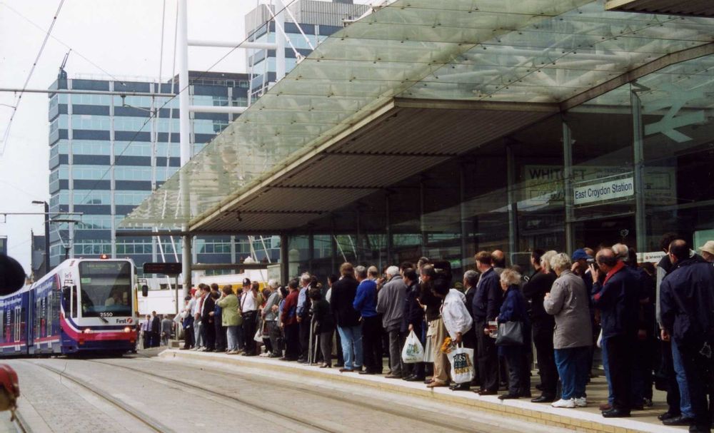 A crowd of people standing on the low tram platform in front of East Croydon station all look towards an approaching tram.