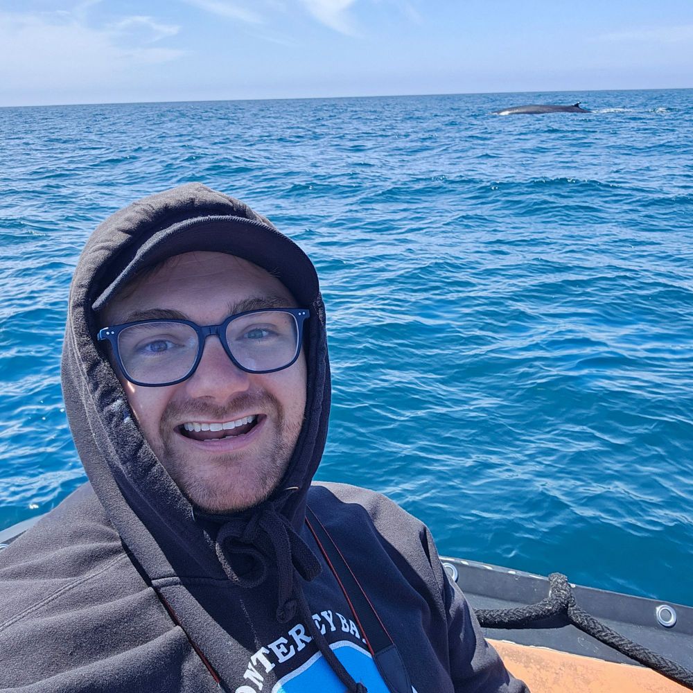 A smiling anthropologist aboard a zodiac vessel off the coast of Southern California. Behind him, a Fin Whale surfaces.