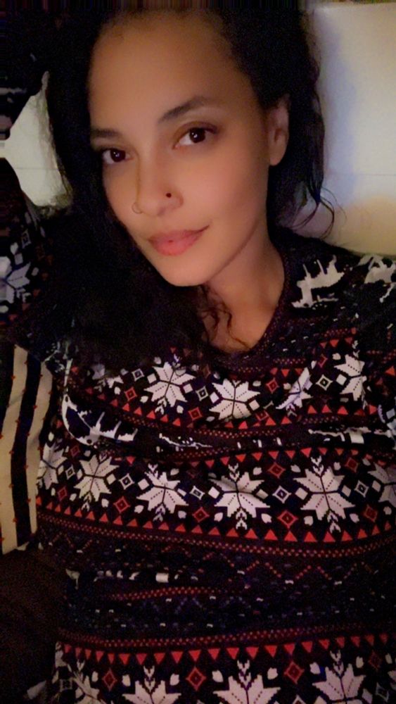 Me with messy curly hair, bare faced with both sides of my nose pierced with black rings. A black long sleeve pajamas holiday shirt on. Laying in bed. 