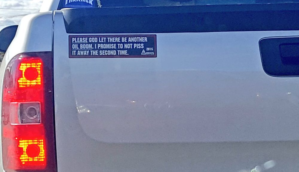 A bumper sticker on the back of a white pickup truck that reads “PLEASE GOD LET THERE BE ANOTHER OIL BOOM. I PROMISE TO NOT PISS IT AWAY THE SECOND TIME.”