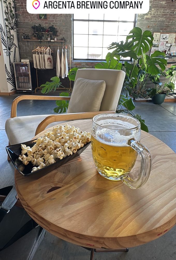Photo of beer in handled mug next to popcorn on small side table. A chair, plans and surroundings in the background.