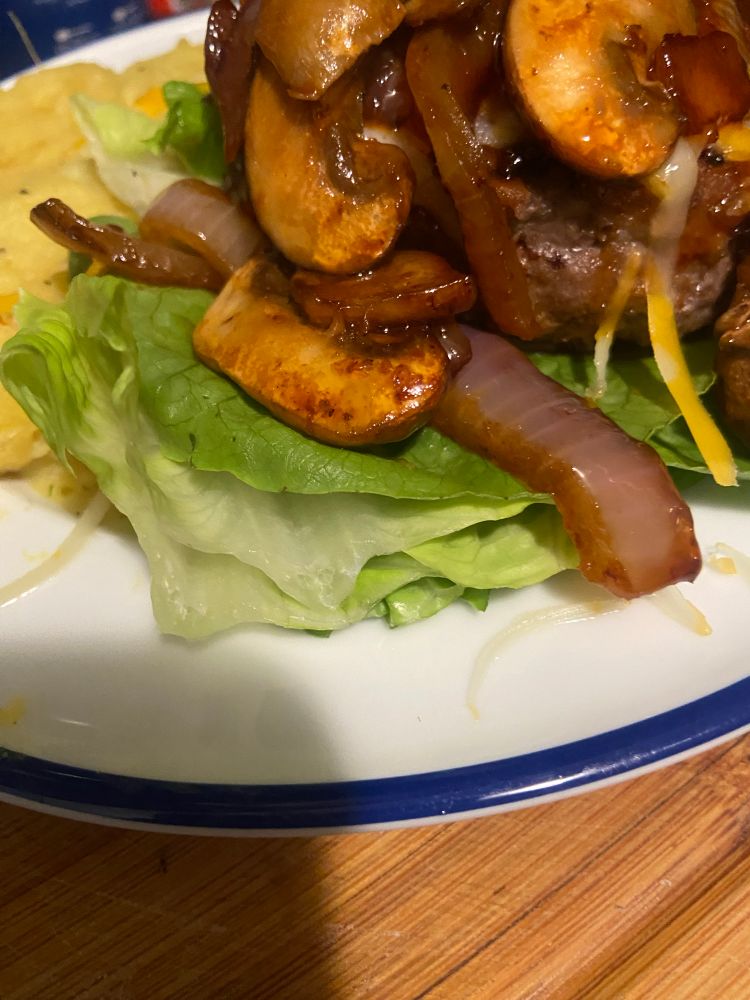 An open face burger with cooked red onions, mushrooms and melted cheese on top of a burger patty. There is mashed potatoes on the side.