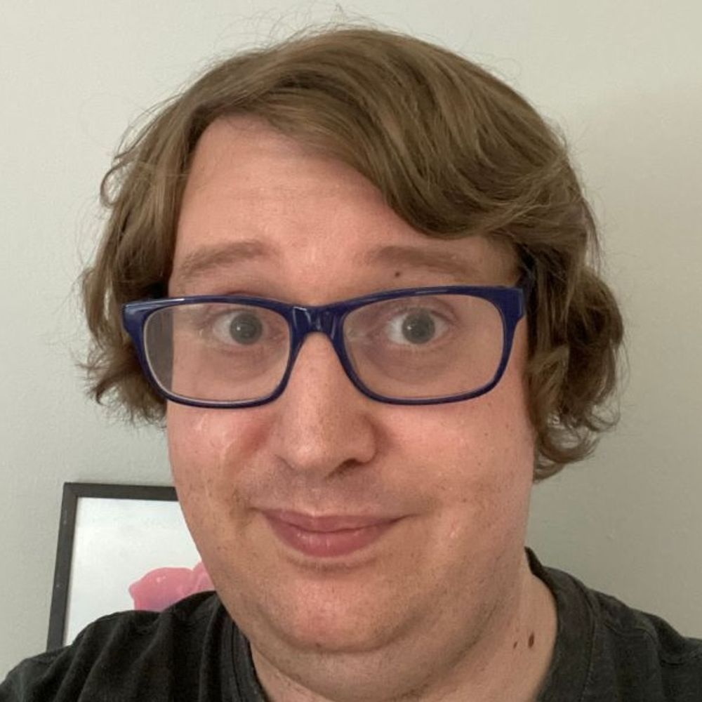A selfie of me cleanly shaven with blue framed glasses and long brown hair with hints of blonde.