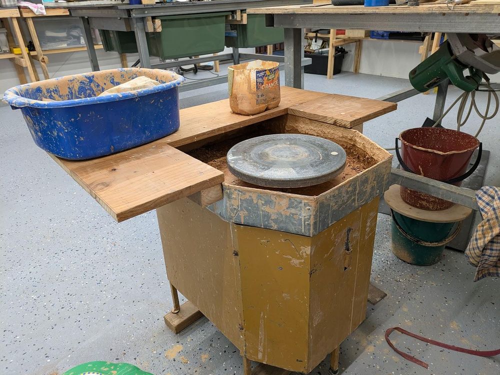 A photo of an old dirty pottery wheel with a wooden shelf built around the top metal splash pan. There is the bottom half of a 3L bottle of orange juice filled with dirty water, and a blue tub spattered with clay mud.