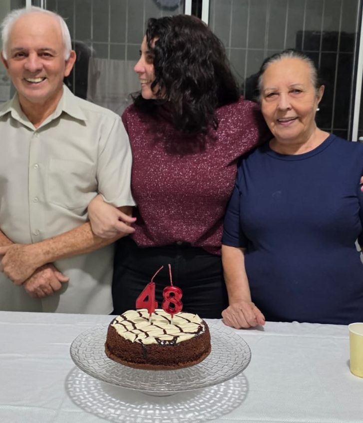 Um bolo de chocolate com cobertura de leite ninho em primeiro plano, atrás, um casal de idosos com a filha mais linda no meio.