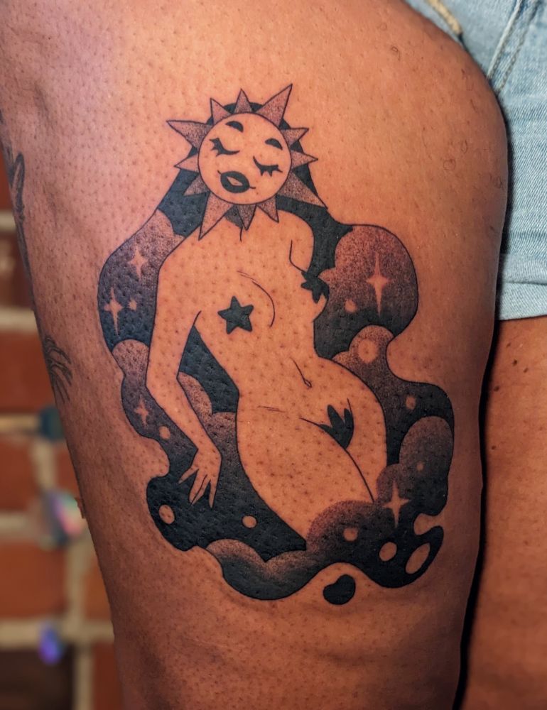 A black and grey tattoo on dark skin. The piece features a nude figure with a sun face and a serene, closed eyes expression. Her hair is made of clouds. 