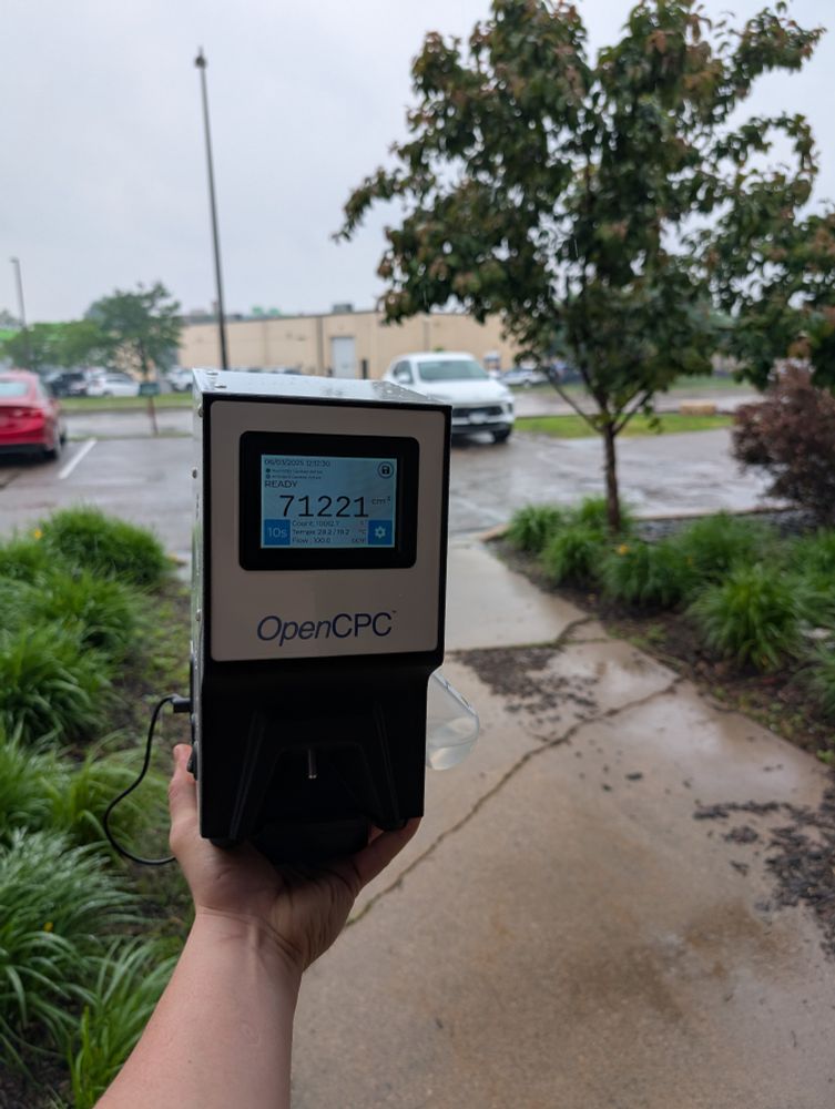 The OpenCPC, a square white and black box displays a particle concentration of 71221 particles/cm^3. The background is a parking lot with shirts and greenery, with a large warehouse in the background. It's overcast and rainy in the picture.