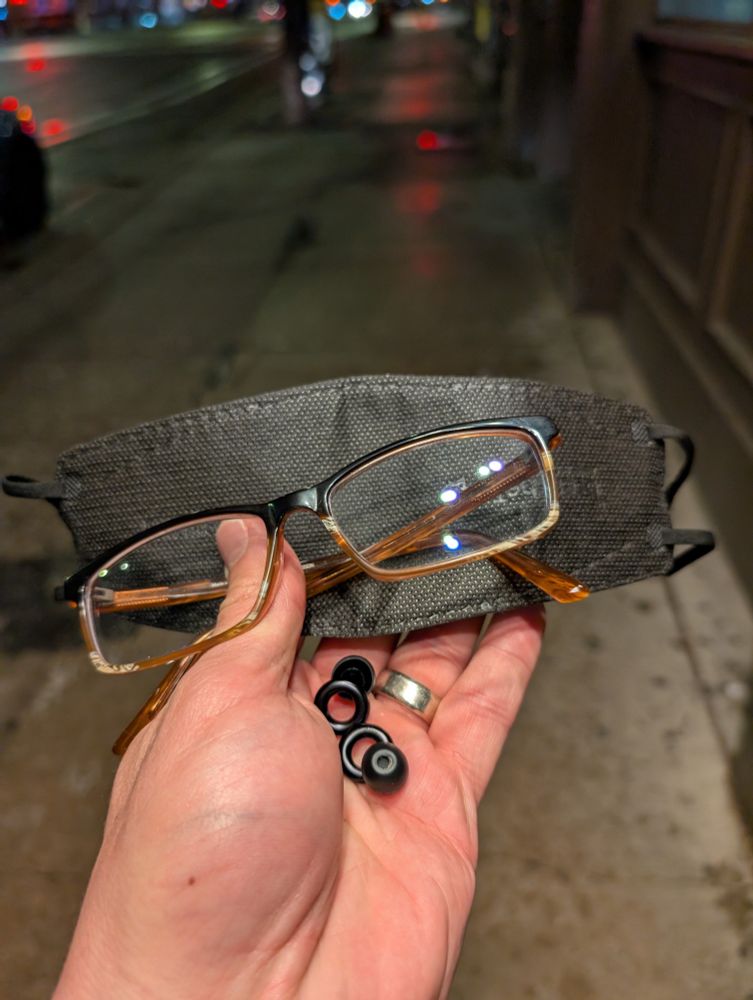 In Aaron's hand is a pair of glasses, ear plugs and black mask with a back drop of a city street.